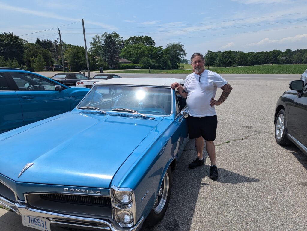 Derek in a white T shirt and black shorts posing beside his blue 1967 Pontiac LeMans, with its white top up. 