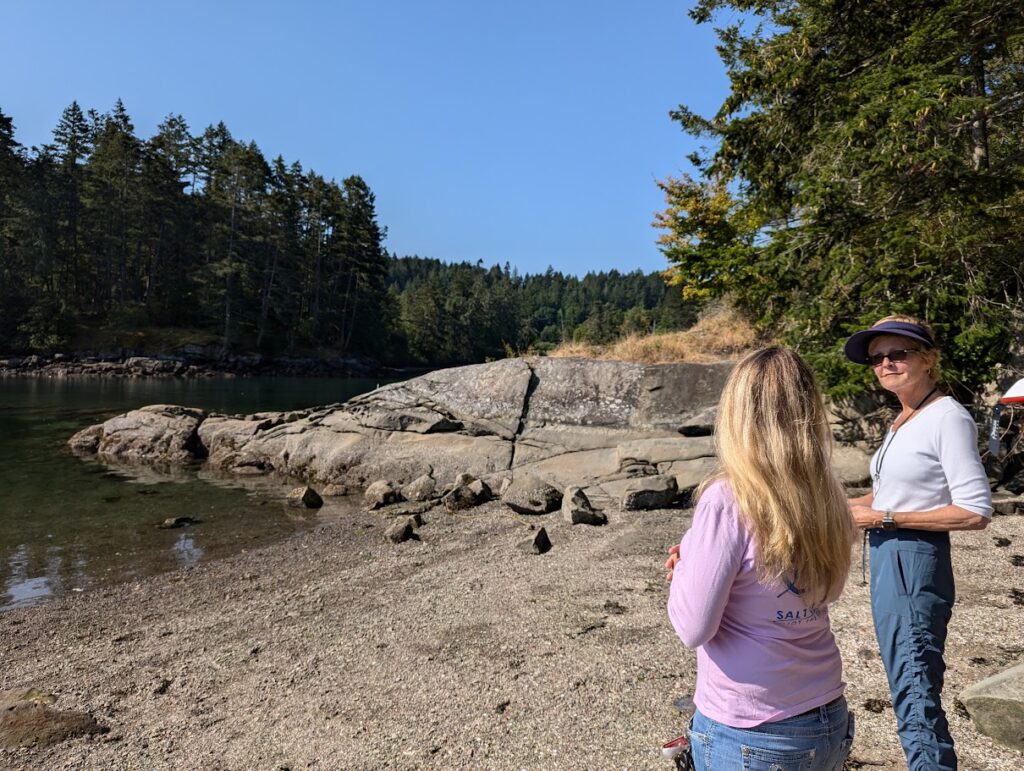 Sarah Smith tells Erin Davis about the island as they stand on a beautiful, sandy shore with rocks, trees, and water in the background. 