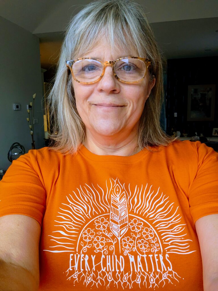 Me in a selfie showing my orange shirt with Every Child Matters and some artwork in white. 