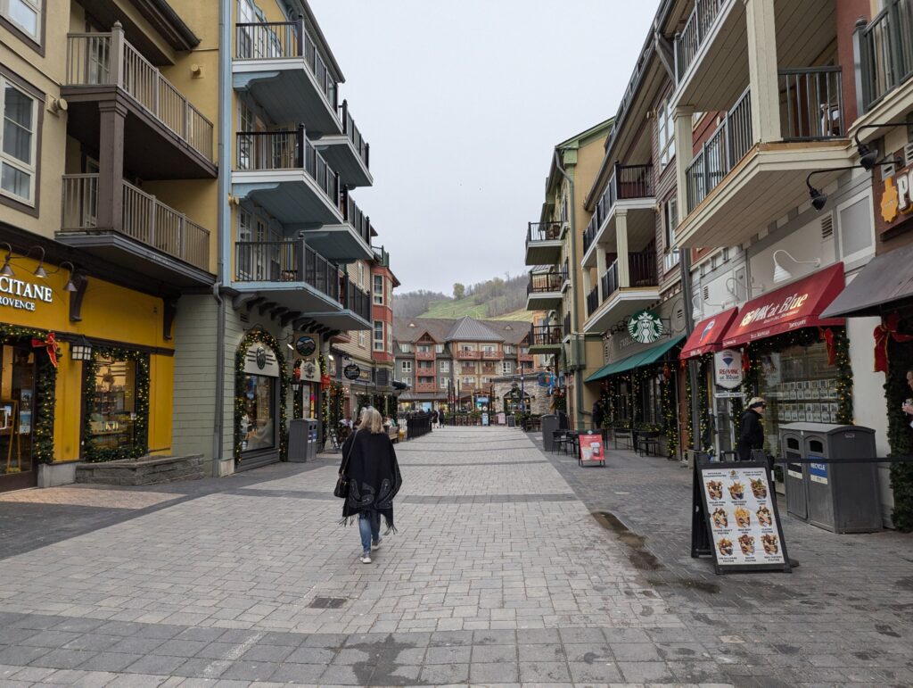 The back of me as I walk down a wide stone walkway with shops on either side. I'm wearing jeans and a heavy black and grey poncho.