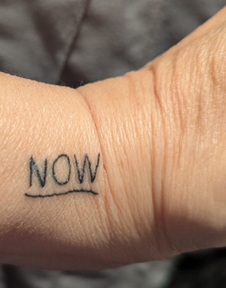 My wrist in closeup with the word NOW in capital letters and a wavy line underneath. 