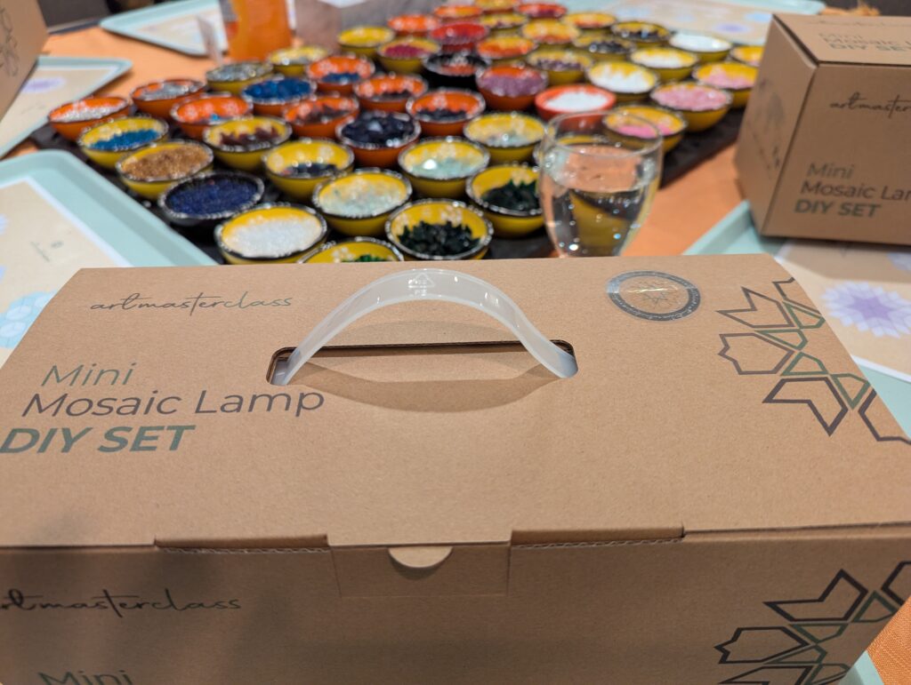 Cardboard box in the foreground has Mini Mosaic Lamp DIY SET printed on it. Dozens of small bowls in the background are filled with different coloured beads and tiles.