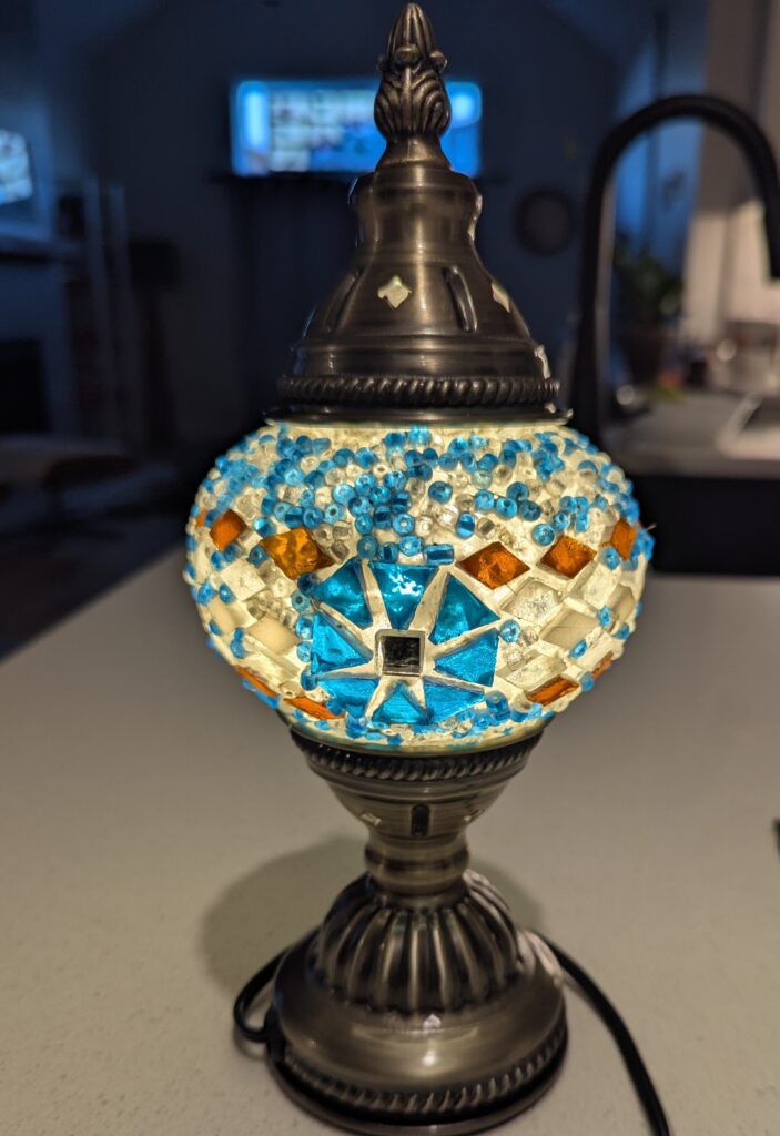 Finished lamp with a burnished brass-looking top and bottom, plugged in, with the blue and copper beads and tiles lit well.