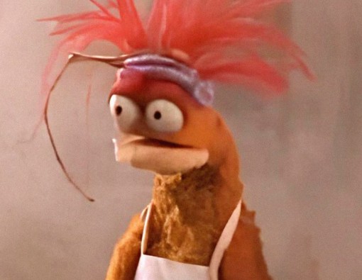 Weird looking muppet-like creature with huge eyes, orange feathers for hair, wearing an apron. 